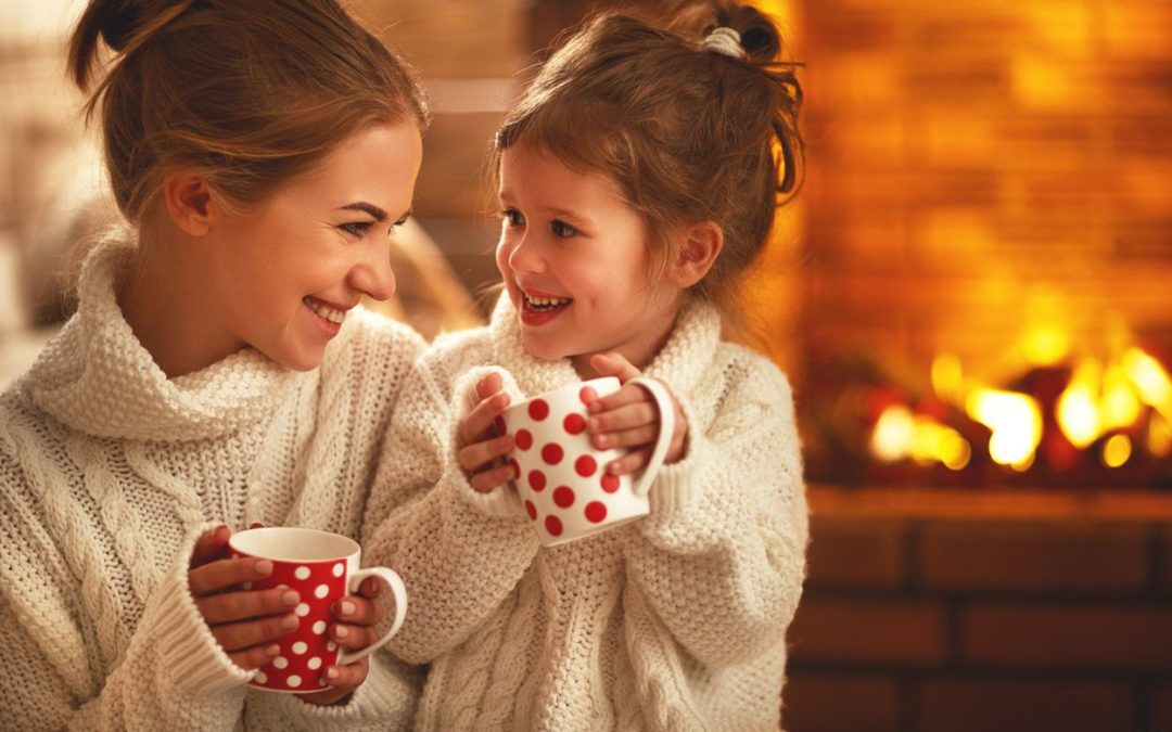 A mother and daughter holding coffee mugs in front of a fireplace.